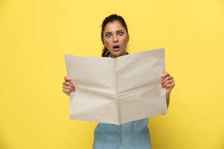 Amazed casual woman reading newspaper and gasping while standing on yellow studio background