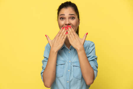 Cheerful casual woman covering her mouth with both hands while standing on yellow studio backgroundの写真素材