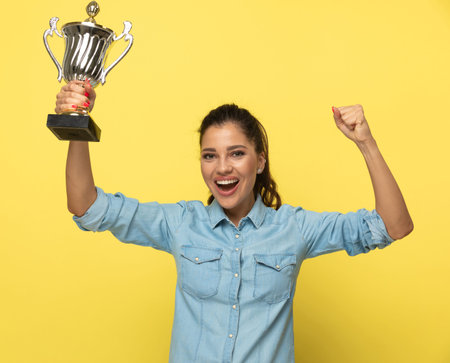 Cheerful casual woman holding trophy and celebrating, shouting while standing on yellow studio...