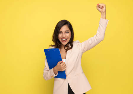 cheerful businesswoman in pink suit holding clipboard, holding fist in the air and celebrating victory on yellow backgroundの写真素材