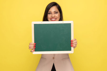 happy young businesswoman in pink suit holding green blackboard and smiling on yellow background