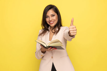 happy young businesswoman in pink suit making thumbs up gesture, reading book and smiling on yellow...