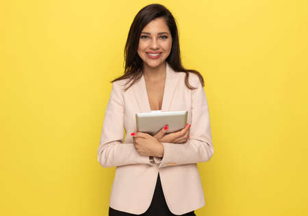 happy young businesswoman in pink suit holding tab and confidently smiling on yellow backgroundの写真素材