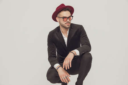 Eager fashion model looking away, wearing hat and sunglasses while crouching on gray studio backgroundの写真素材