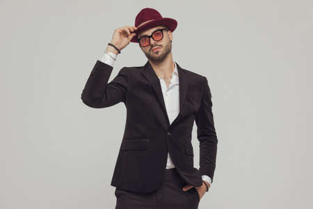 Confident fashion model taking off hat with hand in pocket, wearing sunglasses while standing on gray studio backgroundの写真素材