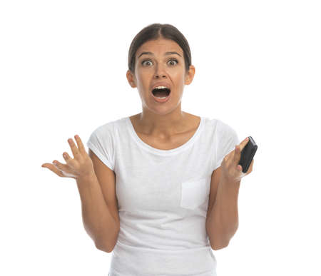 Surprised casual woman gasping and holding phone while standing on white studio backgroundの写真素材