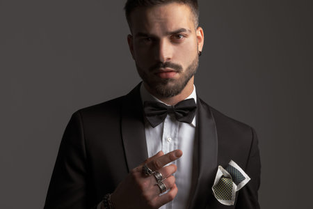portrait of elegant young groom in black tuxedo with bowtie and handkerchief pointing finger and...