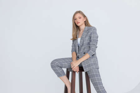 seated elegant businesswoman in gray checkered suit holding hands and confidently posing on light gray background in studioの写真素材