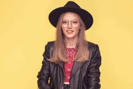 portrait of a young and beautiful casual woman wearing a hat, leather jacket and eyeglasses and smiling against yellow backgroundの写真素材