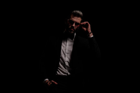 young businessman in black velvet tuxedo fixing sunglasses and holding hand in pocket on black background in studioの写真素材