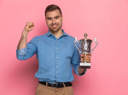 portrait of excited young guy holding silver trophy and celebrating victory, standing and posing on pink background in studioの写真素材