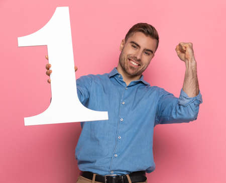 young casual guy in denim shirt holding number one, smiling and holding fists in the air, celebrating and standing on pink background in studioの写真素材