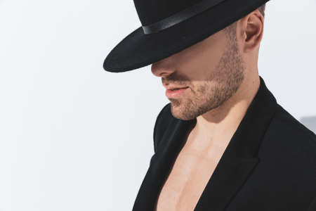 side view of dramatic cool guy in jacket posing shirtless looking down and covering face with black hat on gray background in studioの写真素材