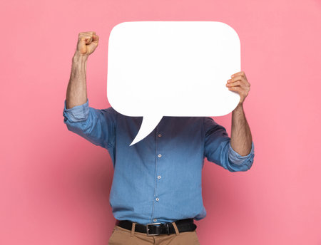 portrait of young man in denim shirt covering face with speech bubble, holding arm up and cheering,...
