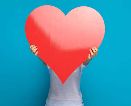 casual woman hiding her face behind a big red heart against blue backgroundの写真素材