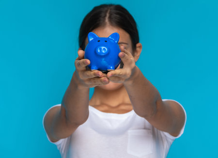casual woman showing her piggy bank at the camera against blue studio backgroundの写真素材