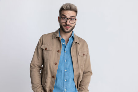 portrait of young casual man in denim shirt with eyeglasses holding hands in pockets and making...