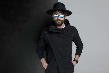 portrait of cool bearded gypsy man wearing sunglasses and black hat holding hand in pockets and confidently posing in studioの写真素材