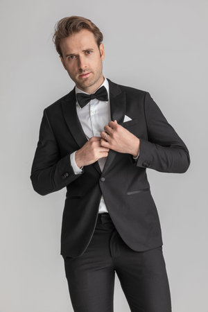 handsome fashion model fixing his tuxedo and looking at the camera on gray backgroundの写真素材