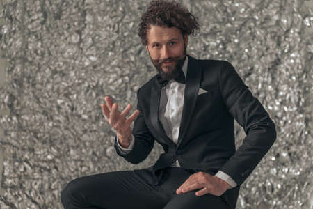 portrait of happy bearded elegant man in black tuxedo holding hand up, gesticulating and inviting while posing on tinfoil background in studioの写真素材