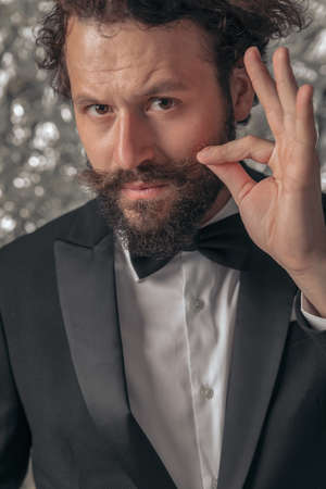 portrait of cool elegant groom in black tuxedo holding hand up and fixing moustache on tinfoil background in studioの写真素材