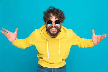 charismatic young guy with retro sunglasses smiling, wearing yellow hoodie and opening arms on blue background in studioの写真素材