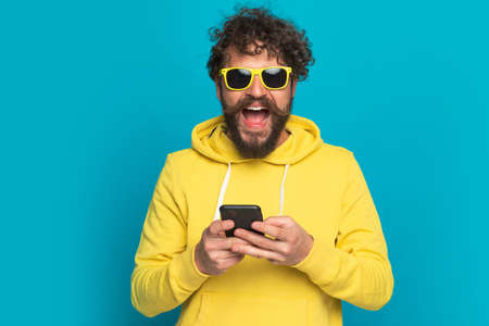 happy young man with sunglasses and yellow hoodie holding phone, reading news and laughing on blue background in studioの写真素材