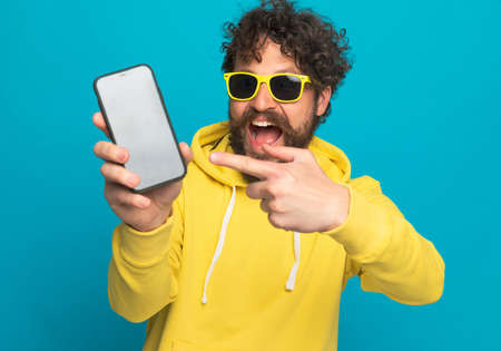 cool unshaved man pointing finger to black empty screen and laughing while posing and standing in front of blue background in studioの写真素材
