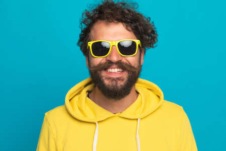 cool young casual man in yellow hoodie wearing sunglasses and smiling on blue background in studioの写真素材