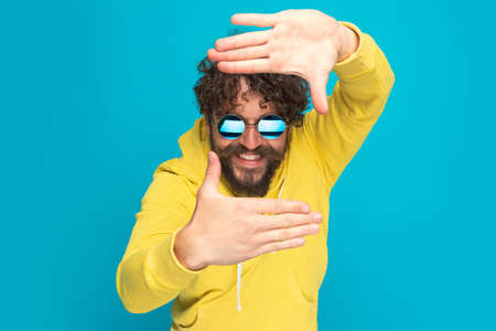 cool unshaved young guy in yellow shirt smiling and holding hands in a framing position while posing on blue background in studioの写真素材