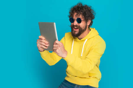 portrait of enthusiastic bearded man in yellow hoodie with retro glasses holding tab, laughing and posing on blue background in studioの写真素材