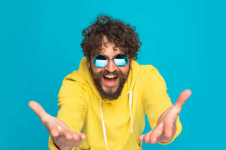 happy young guy in yellow hoodie opening arms, presenting and smiling while standing and posing on blue background in studioの写真素材