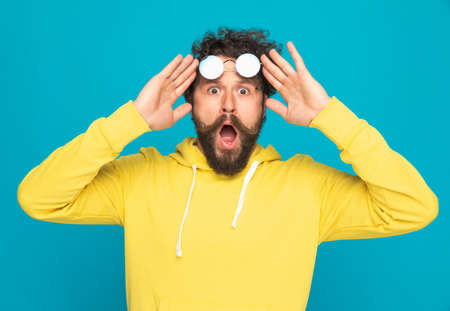 shocked young man holding sunglasses above eyes and opening mouth in a surprised manner on blue background in studioの写真素材