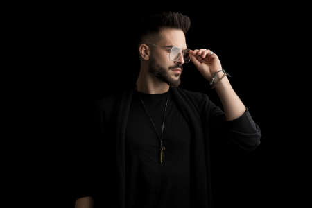cool sexy young man looking to side and arranging eyeglasses while posing in a fashion manner on black background in studioの写真素材