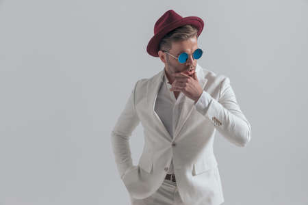 cool fashion model sensually touching his chin, wearing a burgundy hat and sunglasses, holding one hand in pocketの写真素材