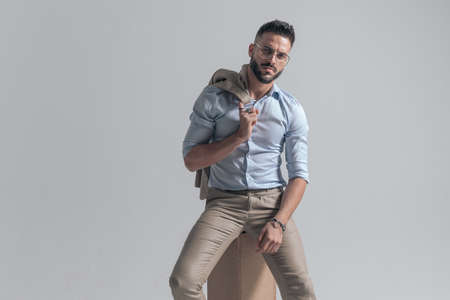 seductive bearded man with jacket over shoulder posing in a fashion way while sitting on grey background in studioの写真素材
