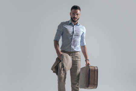 attractive young man with jacket and suitcase posing in a fashion way on grey background in studioの写真素材
