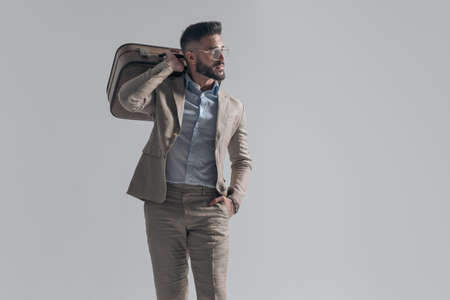 cool businessman with glasses holding hand in pocket and bag over shoulder while looking to side on grey background in studioの写真素材