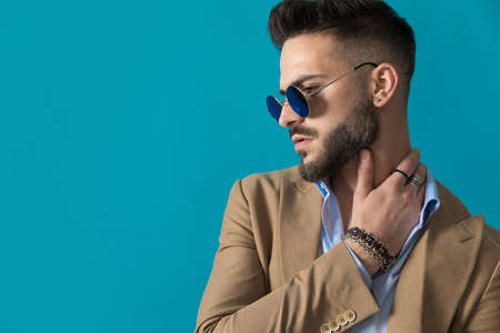side view of sexy young mean with beard wearing retro sunglasses holding hand behind neck and looking to side on blue background in studioの写真素材