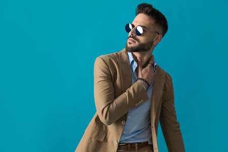 portrait of seductive businessman with retro glasses looking to side and touching neck while posing in a fashion manner in front of blue background in studioの写真素材