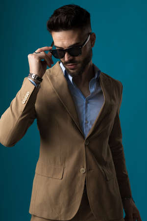 sexy young businessman looking down and adjusting sunglasses while posing on blue background in studioの写真素材