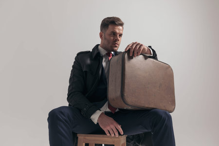 elegant businessman holding suitcase on thigh, looking to side while sitting on grey background in...