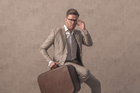 confident young guy arranging glasses and holding suitcase while sitting on beige background in studioの写真素材