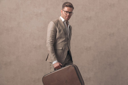 cool young businessman with suitcase walking and being confident on beige background in studioの写真素材