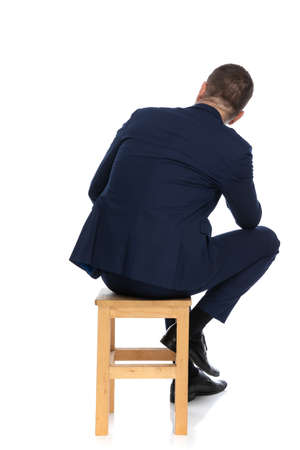 man in elegant suit holding elbow on knee while sitting on wooden chair on white background in studioの写真素材