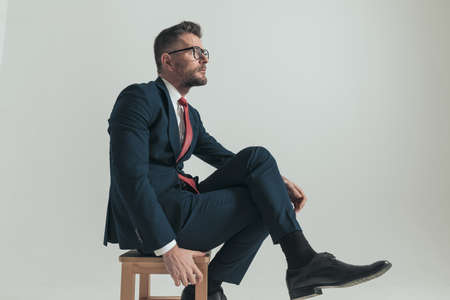 side view of sexy businessman with legs crossed looking up side and thinking while sitting on grey background in studioの写真素材