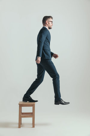 side view of standing businessman in suit stepping on wooden chair and never looking back on grey...