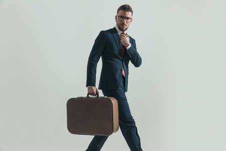 side view of bearded man with glasses adjusting tie, holding luggage and travelling while walking away on grey backgroundの写真素材