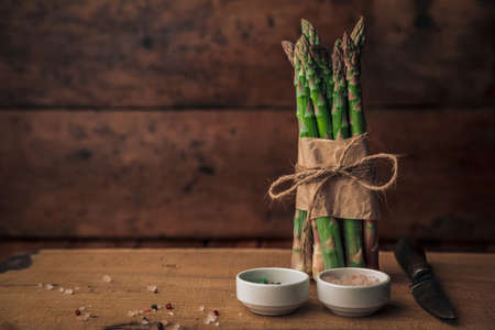 rustic picture with a fresh bunch of asparagus tied with rope on top of a wooden chopping board with salt, pepper and a knife next to itの写真素材