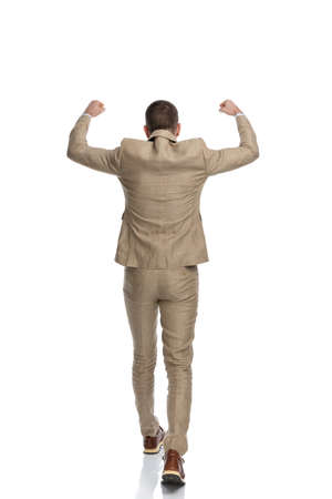 back view of businessman in beige suit holding arms in the air and walking while celebrating victory on white background in studioの写真素材